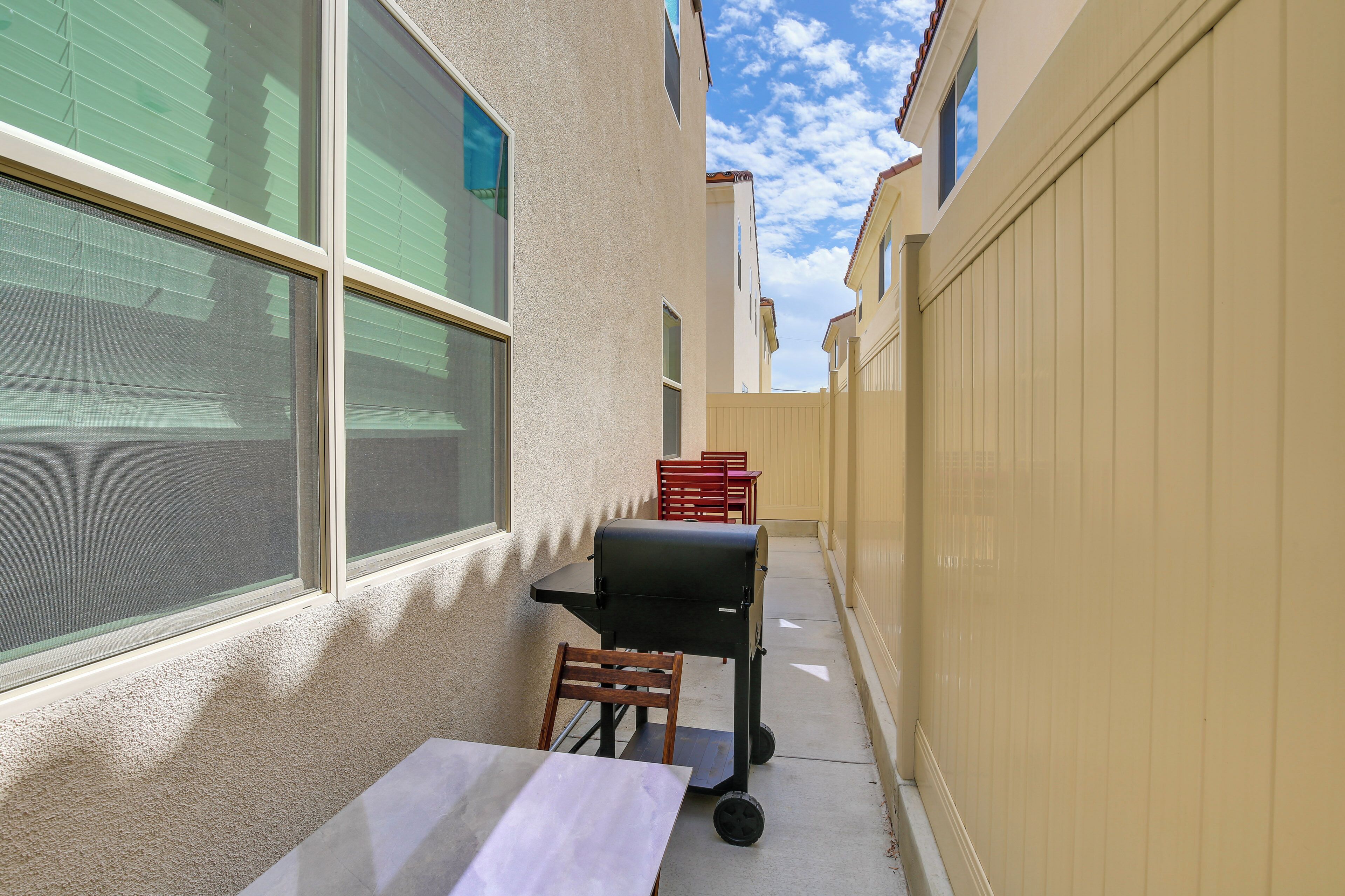 A narrow outdoor patio with a table and chairs, a grill, and a window with blinds.