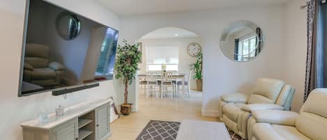 A modern living room with a flat-screen TV, a sofa, a coffee table, and a dining area in the background.