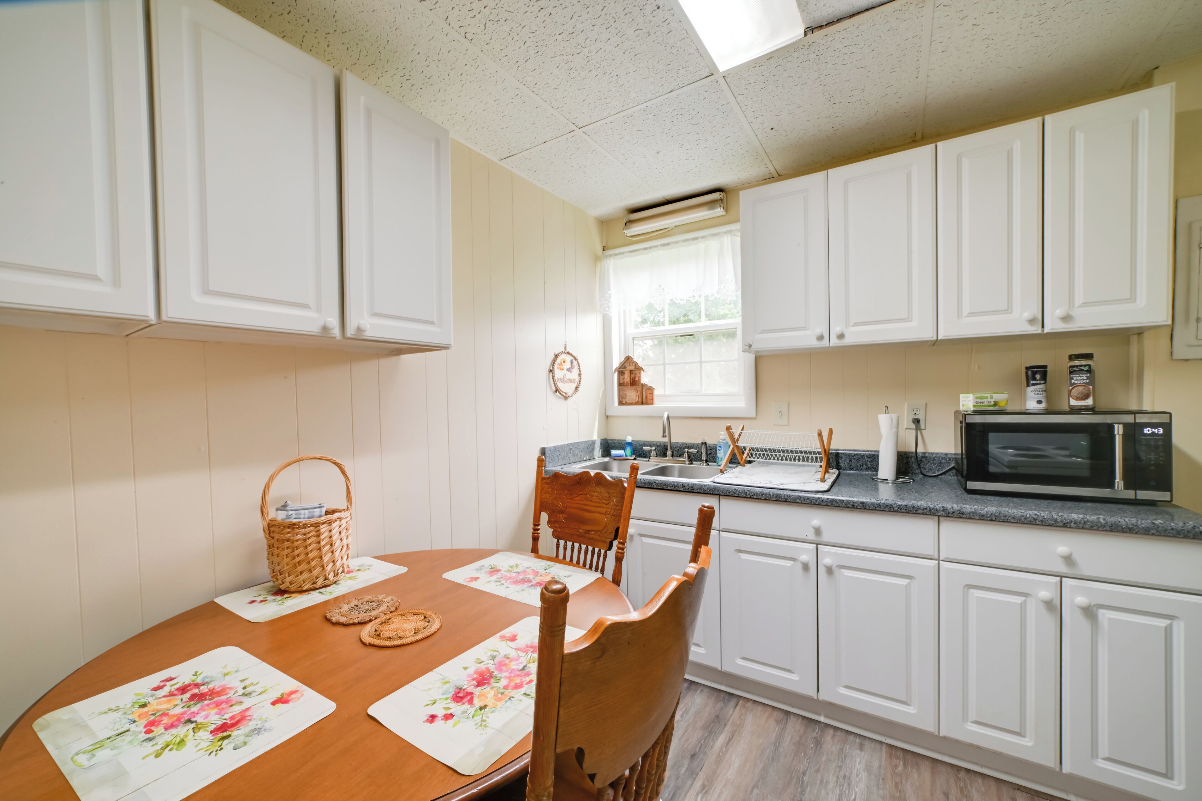 A kitchen with white cabinets, a wooden table with floral placemats, a wicker basket, and a microwave on the counter.