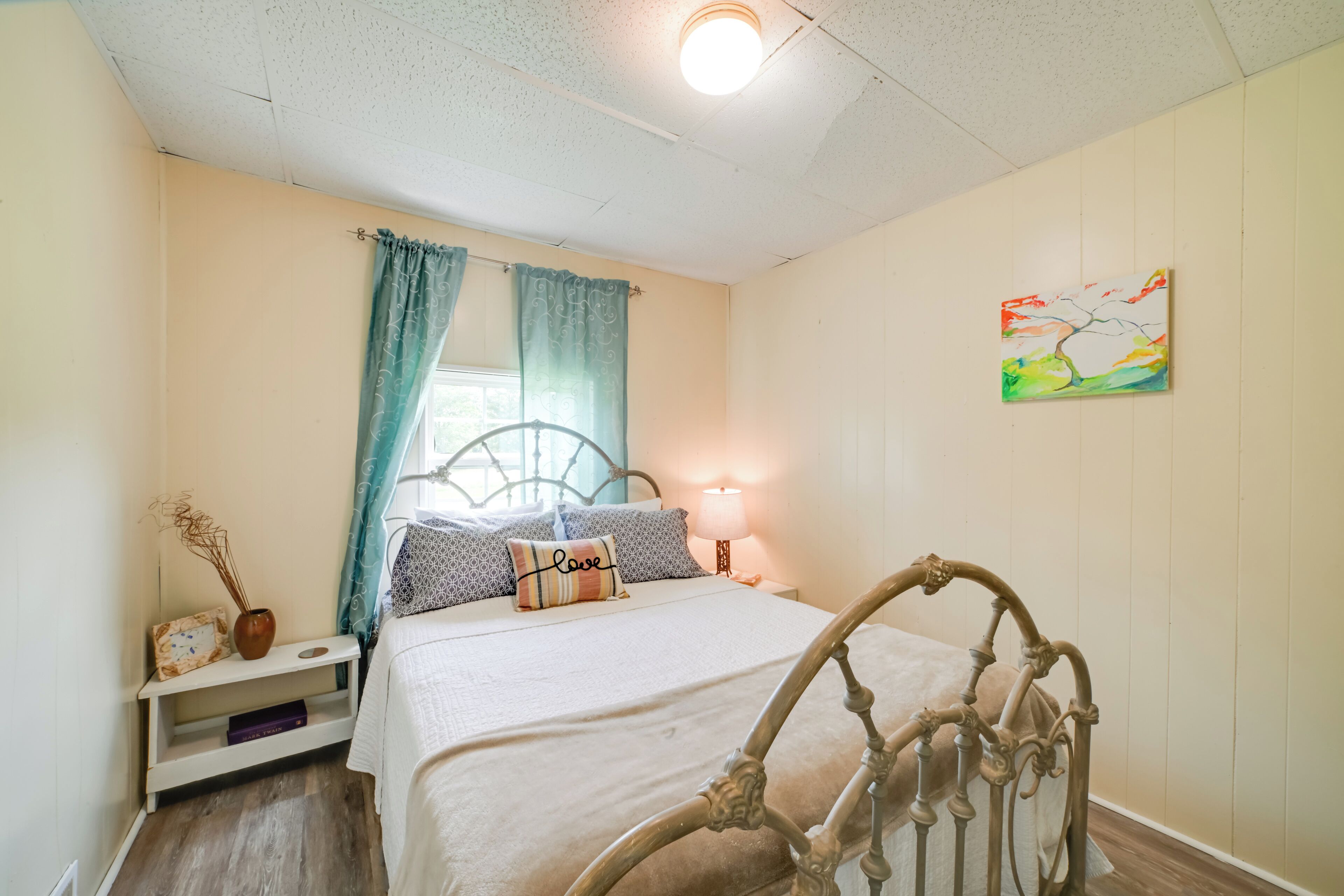 Une chambre à coucher avec un lit, des rideaux, une table de chevet, une lampe et un tableau au mur.