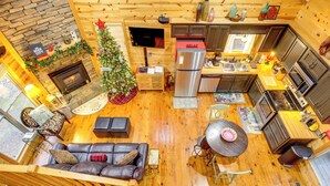 Un acogedor interior de cabaña con chimenea, un árbol de Navidad, una zona de cocina y una sala de estar con sofá de cuero.