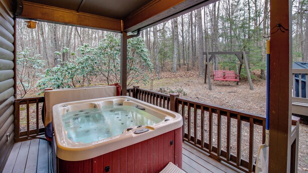 Una terraza de madera con un jacuzzi, un conjunto de columpios de madera y un banco en un área boscosa.