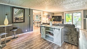 A modern living room with a gray sofa, a coffee table, and a TV. There is a kitchen area in the background with a dining table and chairs. - Riverfront Home w/ Dock ~ 6 Mi to Cheboygan! (Cheboygan)