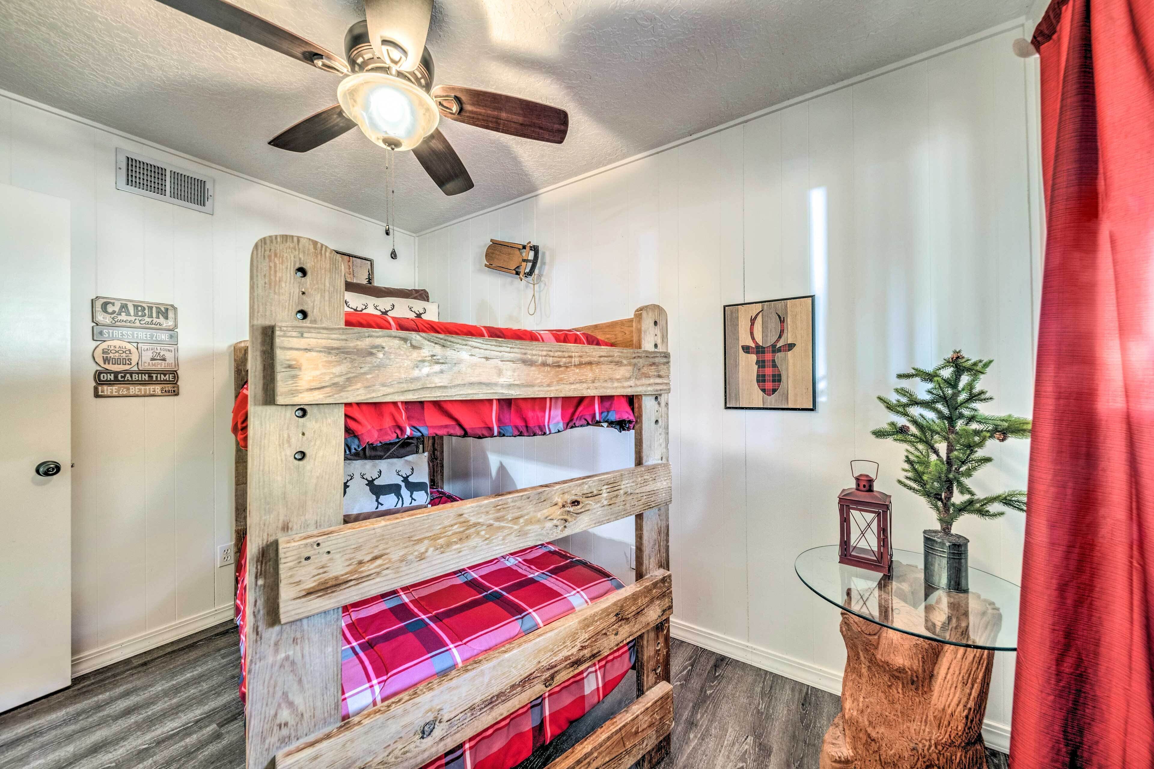 Un lit superposé avec une structure en bois et une literie rouge, un ventilateur de plafond, une petite table avec une plante et un panneau accroché au mur.