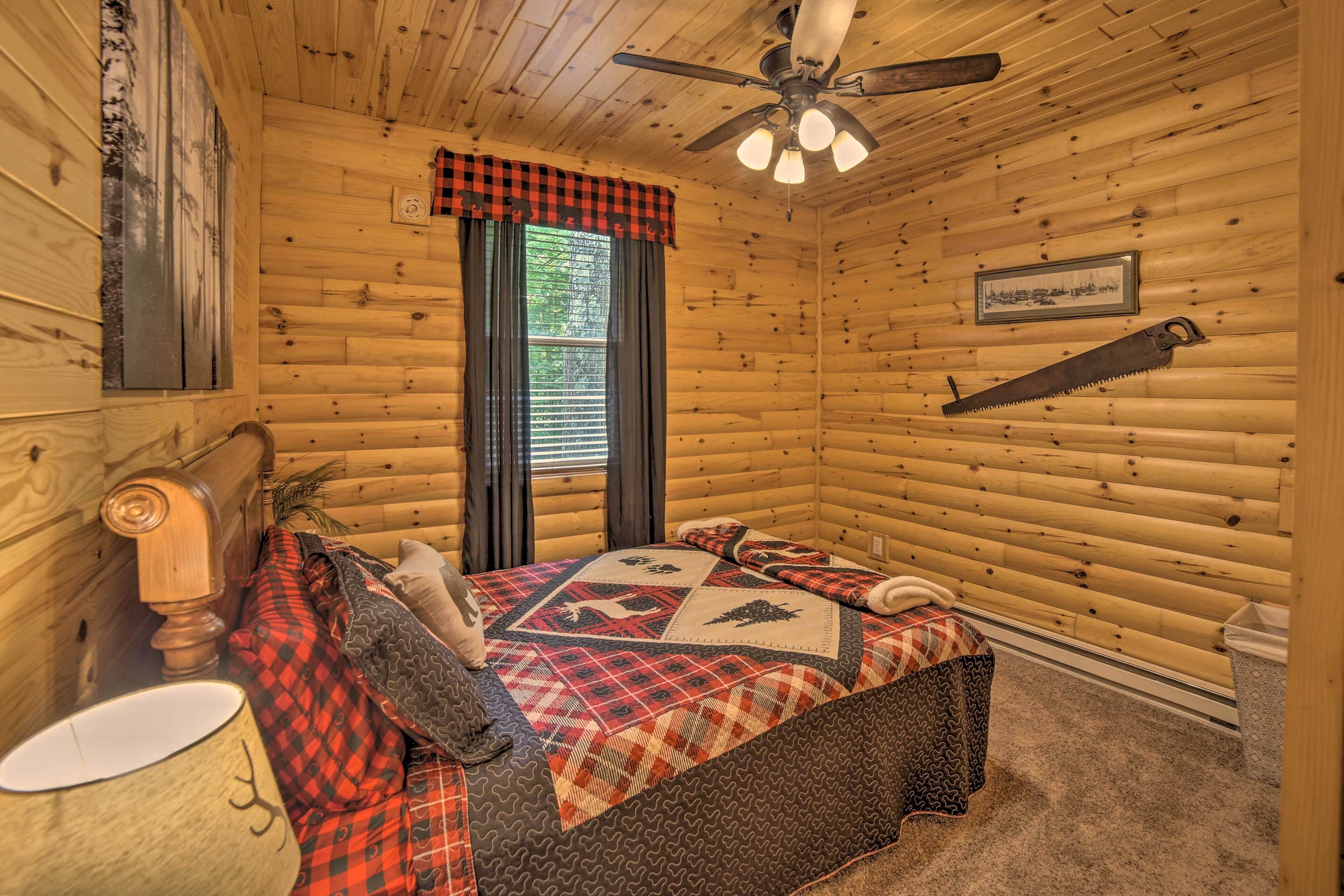 A log cabin bedroom with a bed, a ceiling fan, a window with curtains, and a wall-mounted saw.