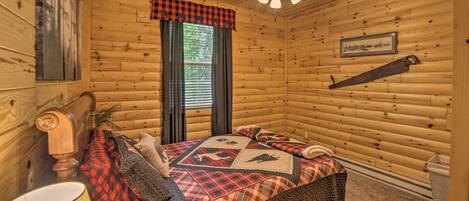 A log cabin bedroom with a bed, a ceiling fan, a window with curtains, and a wall-mounted saw.