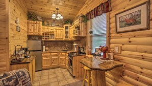 A log cabin kitchen with wooden cabinets, a central island, a refrigerator, and a dining area. - Updated Manistique Log Cabin, Yard & Fire Pit (Manistique)