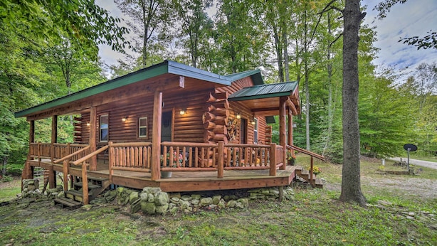 A log cabin with a porch, stone foundation, and a view of the surrounding forest. - Updated Manistique Log Cabin, Yard & Fire Pit (Manistique)