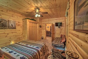 A cozy log cabin room with a bed, a small sofa, a ceiling fan, and a television. - Updated Manistique Log Cabin, Yard & Fire Pit (Manistique)