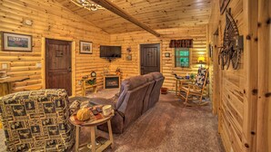 A log cabin interior with a sofa, a patterned armchair, a small table with a pumpkin, and a television on the wall. - Updated Manistique Log Cabin, Yard & Fire Pit (Manistique)