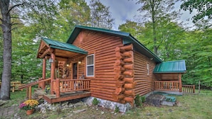 A rustic log cabin with a porch, a small deck, and a stone foundation. - Updated Manistique Log Cabin, Yard & Fire Pit (Manistique)