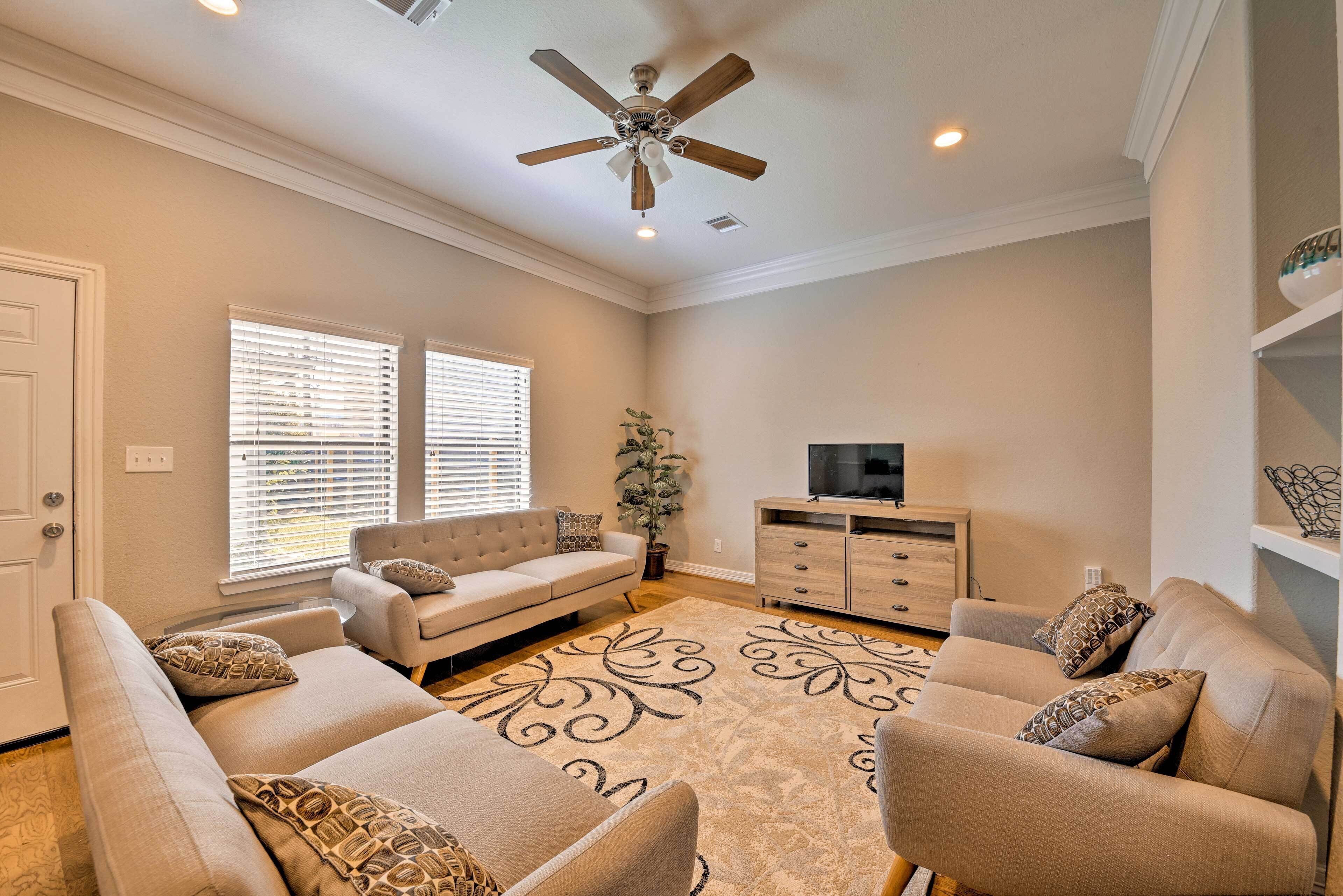 A living room with a beige sofa set, a flat-screen TV, a ceiling fan, and a decorative rug.