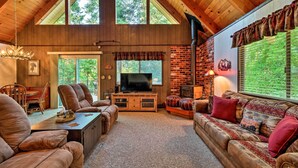 A spacious living room with a high ceiling, wooden beams, a fireplace, and a large window overlooking greenery. - Twain Harte Mountain Cabin w/ Deck - 3 Mi to Dtwn (Twain Harte)
