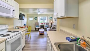 A kitchen with white cabinets, a microwave, a stove, and a sink. There is a dining area with chairs and a table, and a window with curtains. - Winchester Bay Apt Near Dunes & State Parks! (Reedsport)
