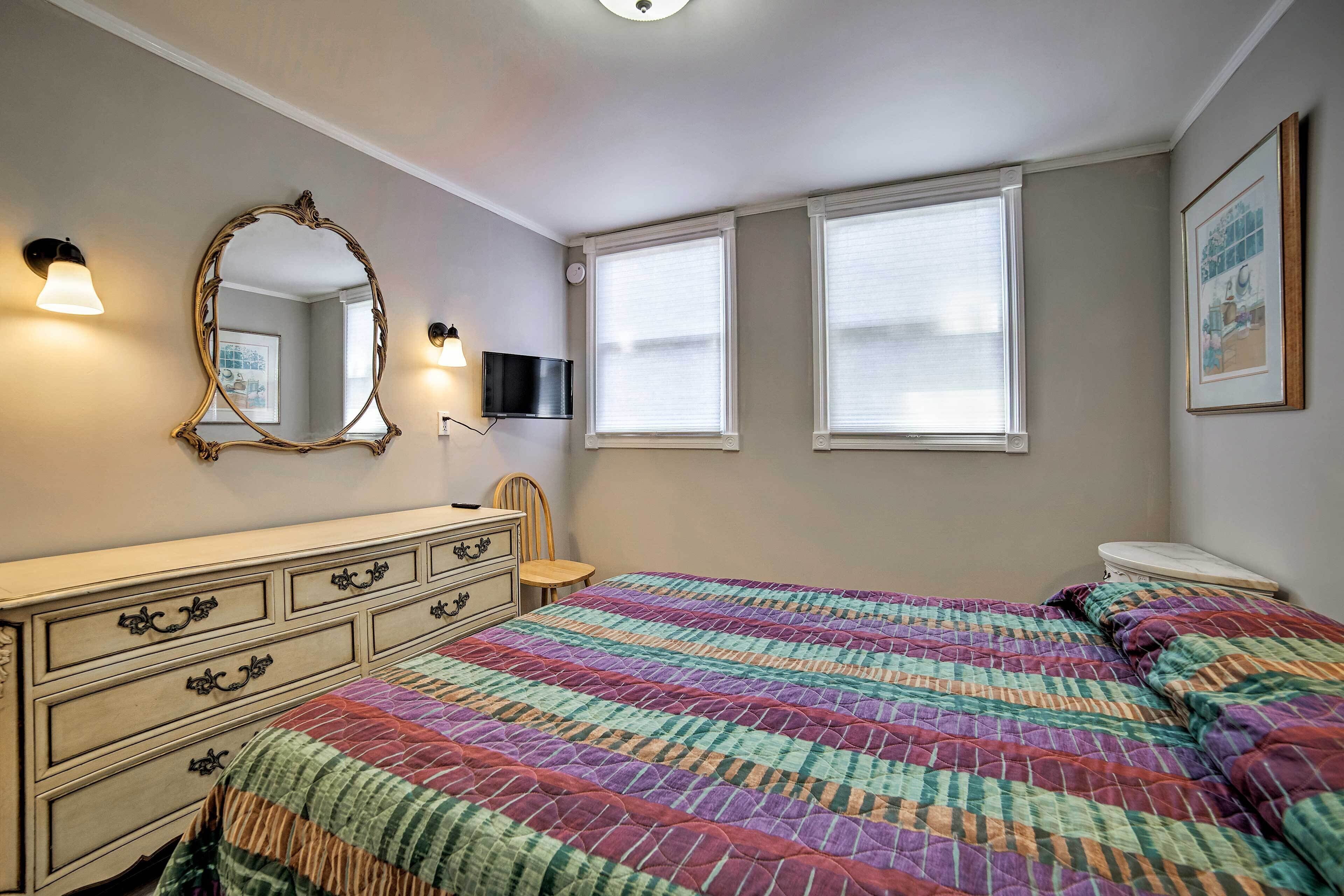 A bedroom with a large bed, a dresser, a mirror, and two windows with blinds.