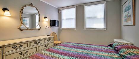 A bedroom with a large bed, a dresser, a mirror, and two windows with blinds.