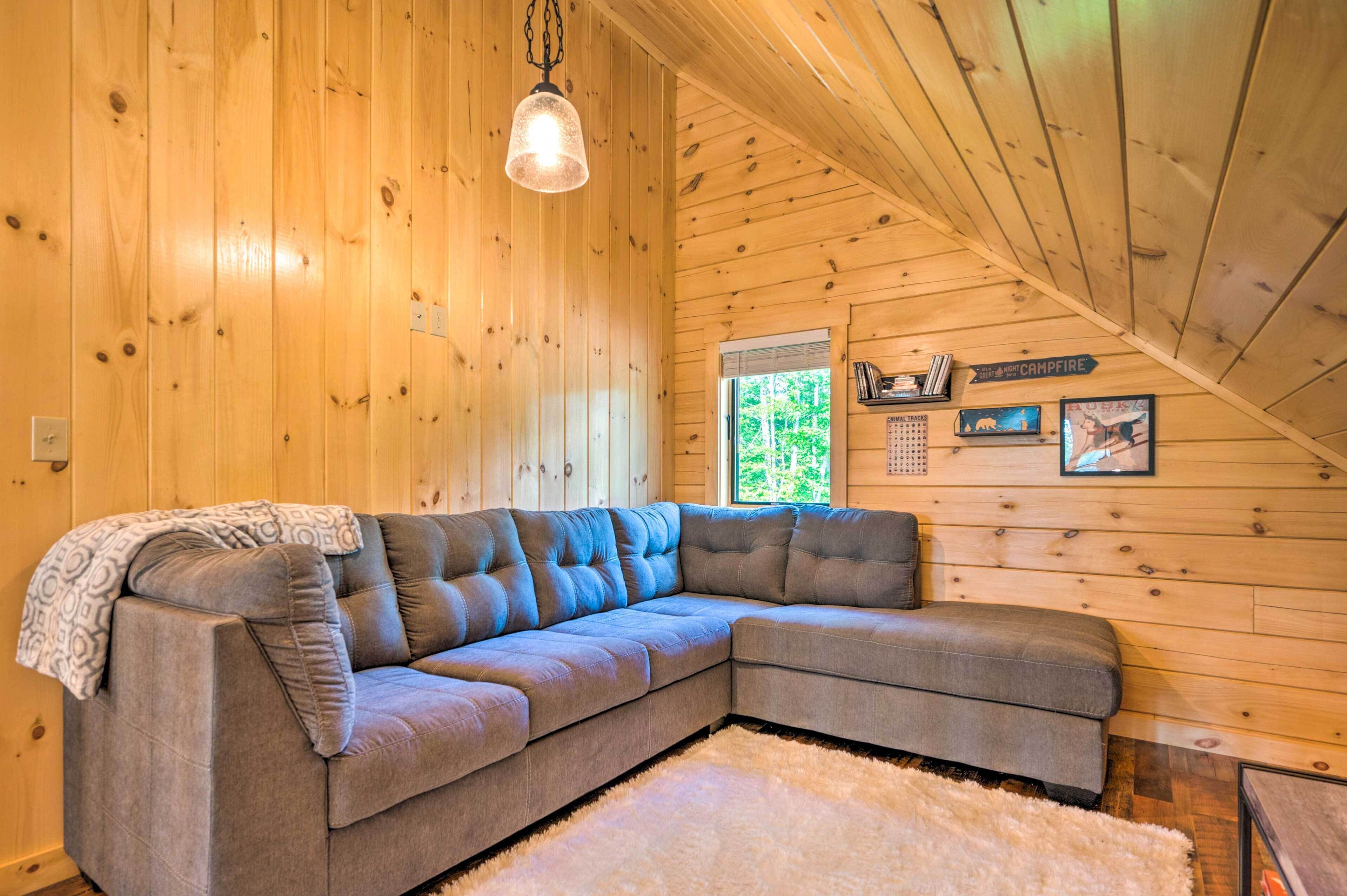 Un cuarto acogedor con sofá modular, paredes de madera, lámpara colgante y ventana con vista a follaje.