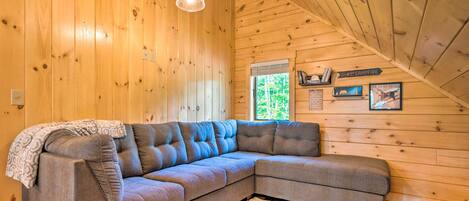 Un cuarto acogedor con sofá modular, paredes de madera, lámpara colgante y ventana con vista a follaje.
