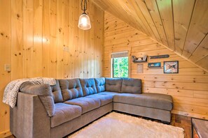 A cozy room with a sectional sofa, wooden walls, a hanging light fixture, and a window with a view of greenery. - Peaceful North Haverhill Haven w/ Wood Fire Pit (North Haverhill)