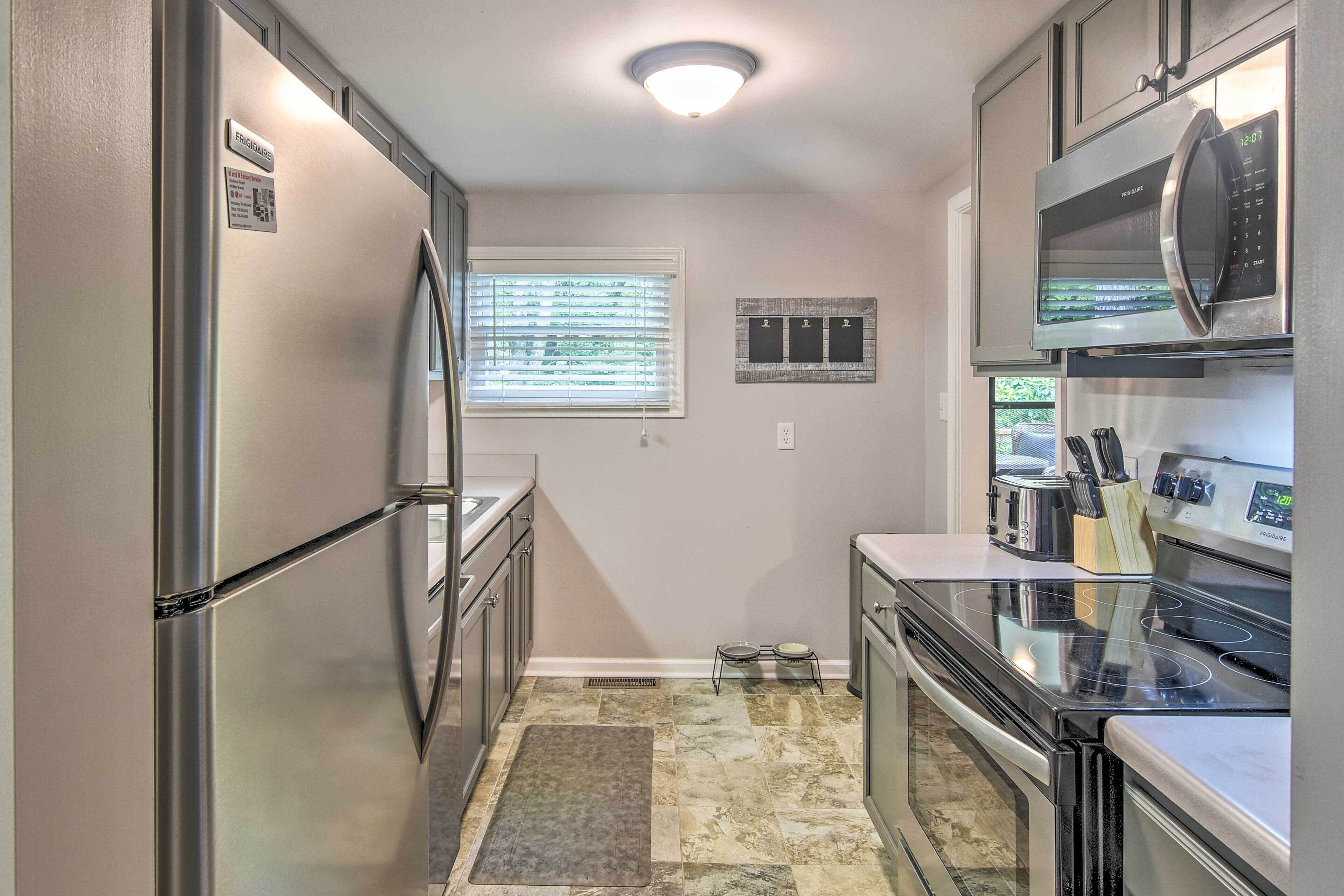 A compact kitchen with stainless steel appliances, a window with blinds, a pet feeding station, and a tiled floor.