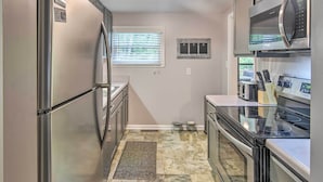 A compact kitchen with stainless steel appliances, a window with blinds, a pet feeding station, and a tiled floor.