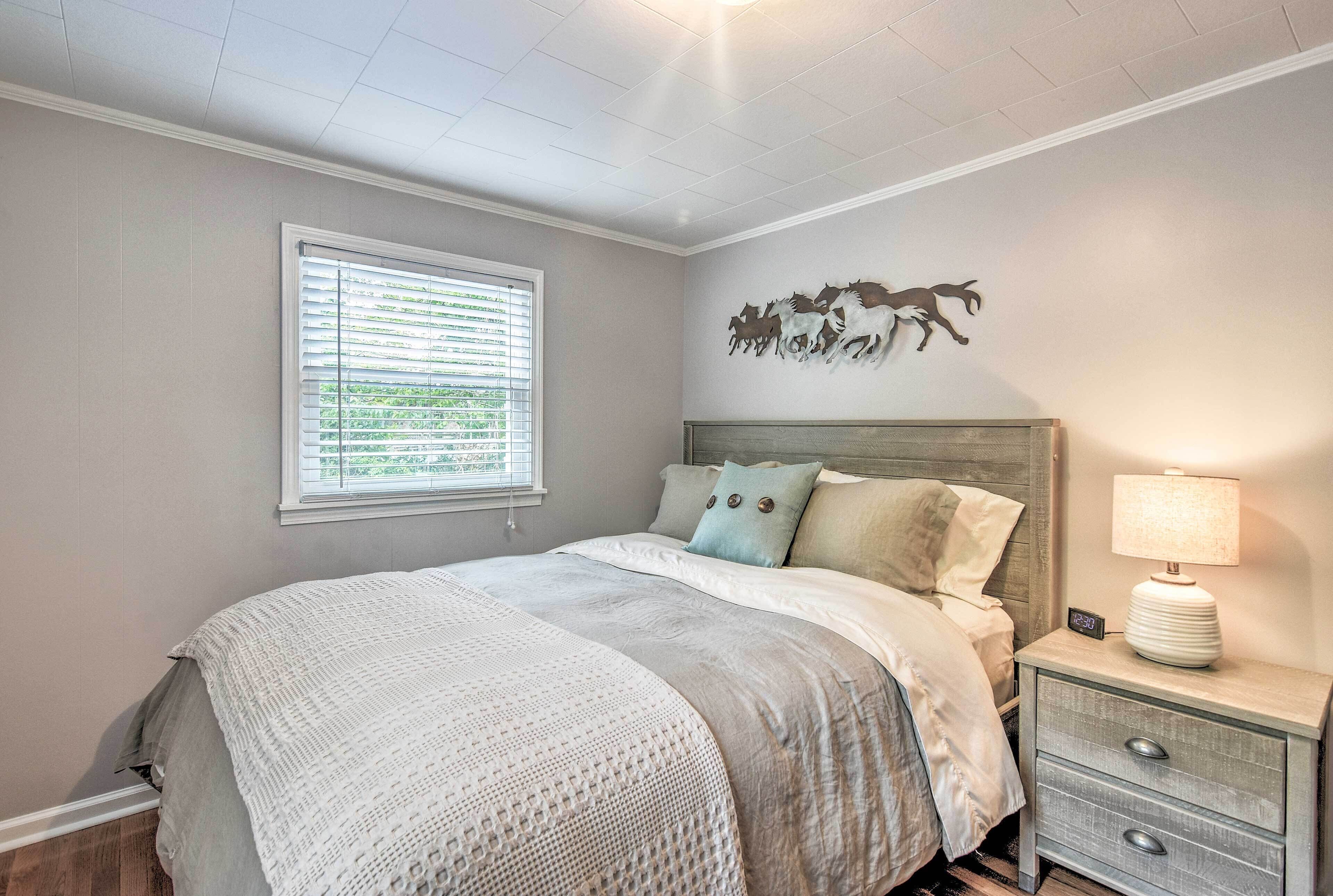 A bedroom with a bed, a nightstand with a lamp, a wall clock, and a window with blinds.