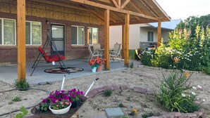 Un patio cubierto con mobiliario exterior, un carretillón con flores, y un jardín con diversas plantas y flores.