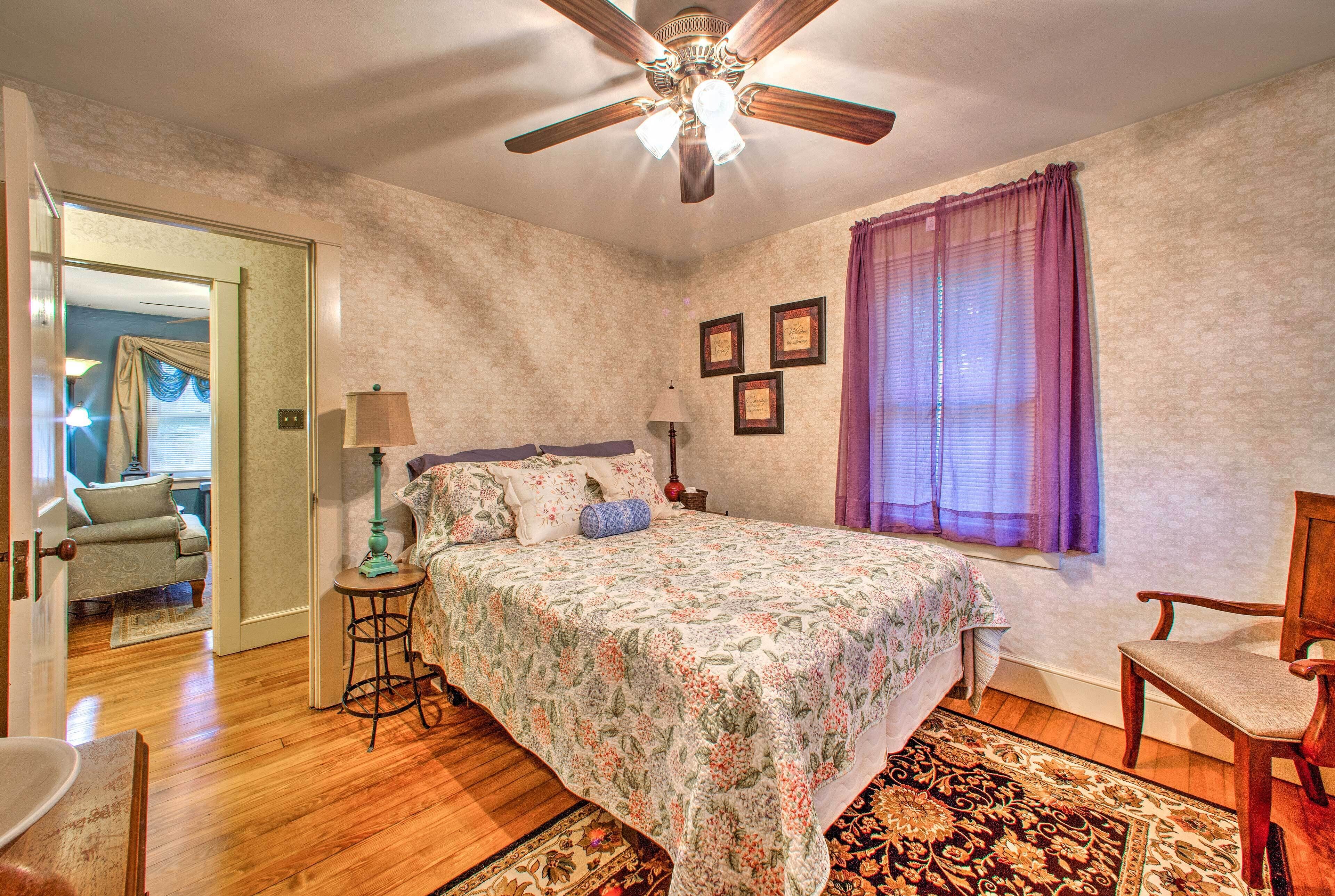 Une chambre à coucher avec un lit, un ventilateur de plafond, une fenêtre avec des rideaux violets et une chaise.