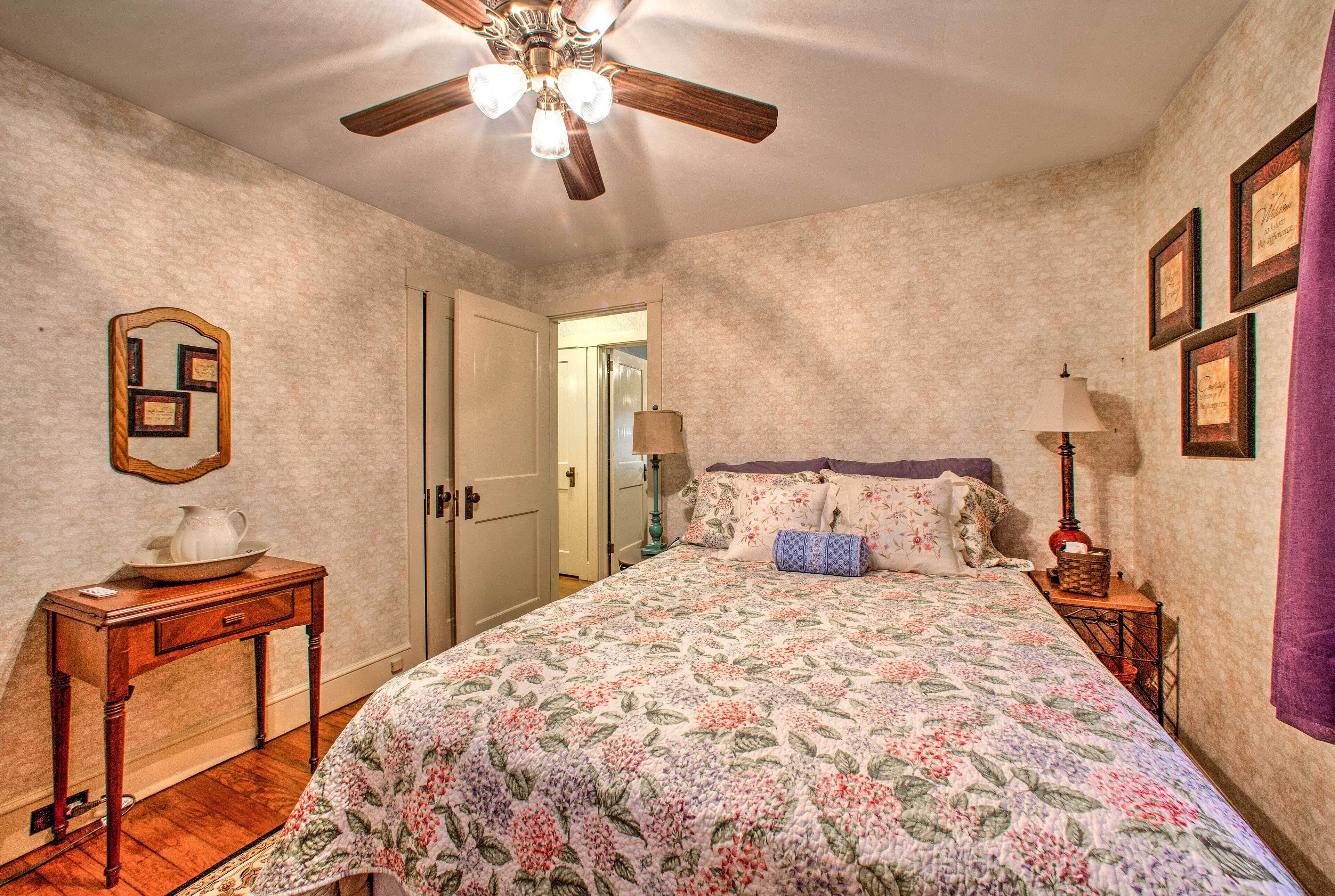 A bedroom with a floral bedspread, a wooden nightstand, a ceiling fan, and framed pictures on the wall.