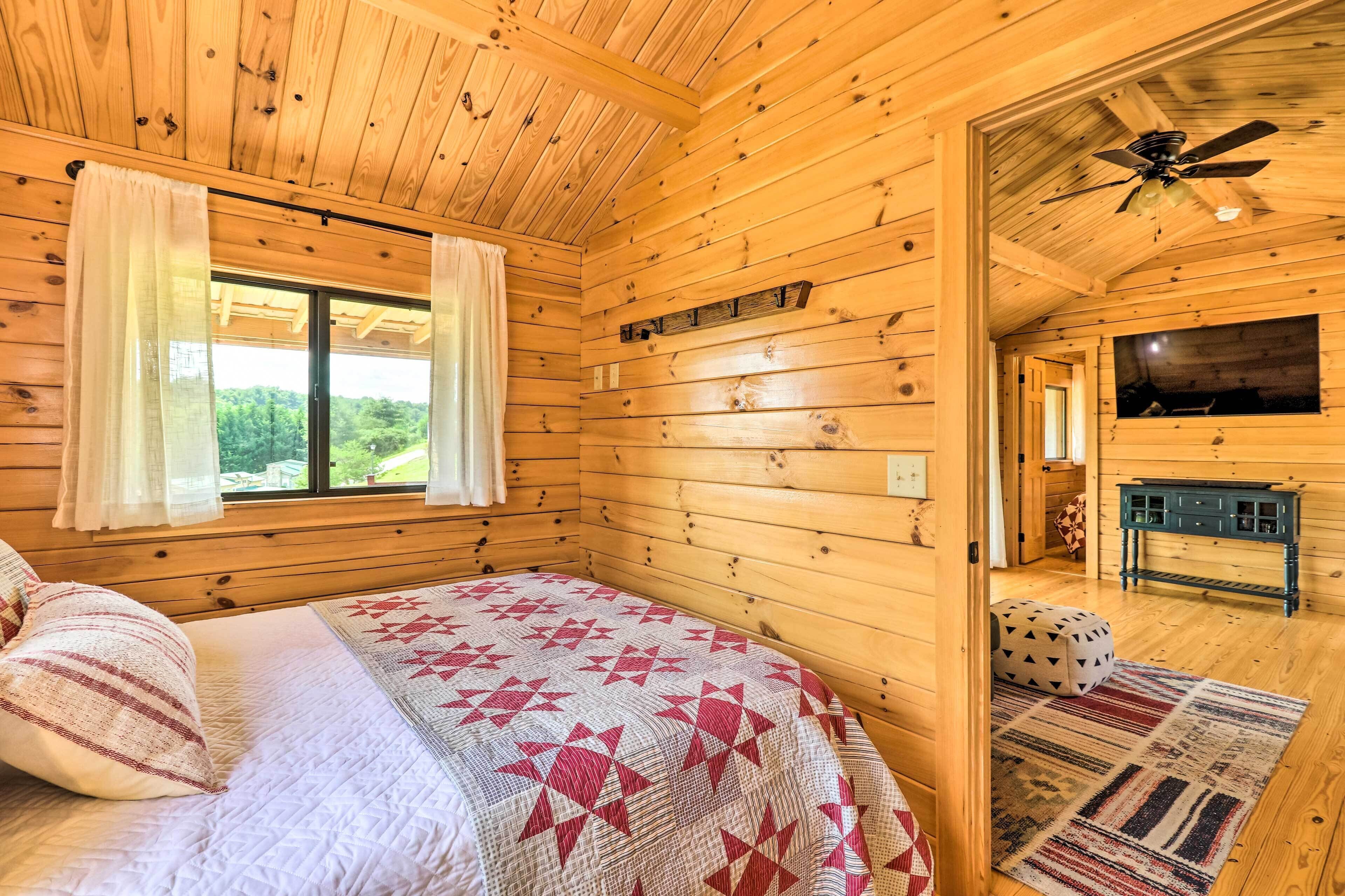 A wooden cabin bedroom with a bed, curtains, a ceiling fan, a TV, and a fireplace.