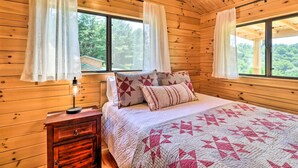 A wooden cabin bedroom with a bed, curtains, a nightstand, and a window. - Grill & River Access: Tiny Cabin in Sparta! (Sparta)