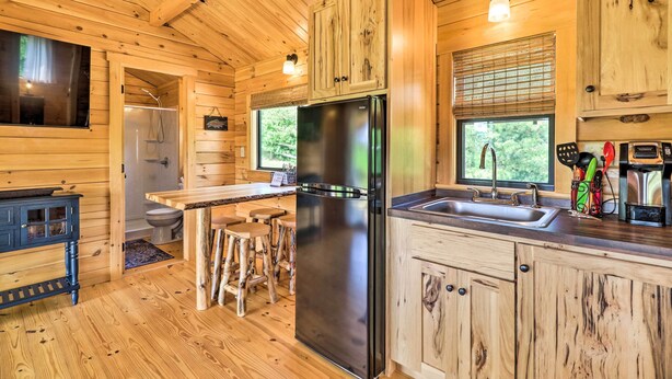 A wooden cabin interior with a kitchen area featuring a black refrigerator, wooden cabinets, and a sink. - Grill & River Access: Tiny Cabin in Sparta! (Sparta)