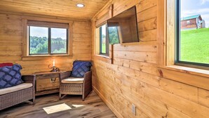 A wooden cabin interior with a sofa, a small table, and a chair. There are two windows providing views of the outdoors. - Grill & River Access: Tiny Cabin in Sparta! (Sparta)