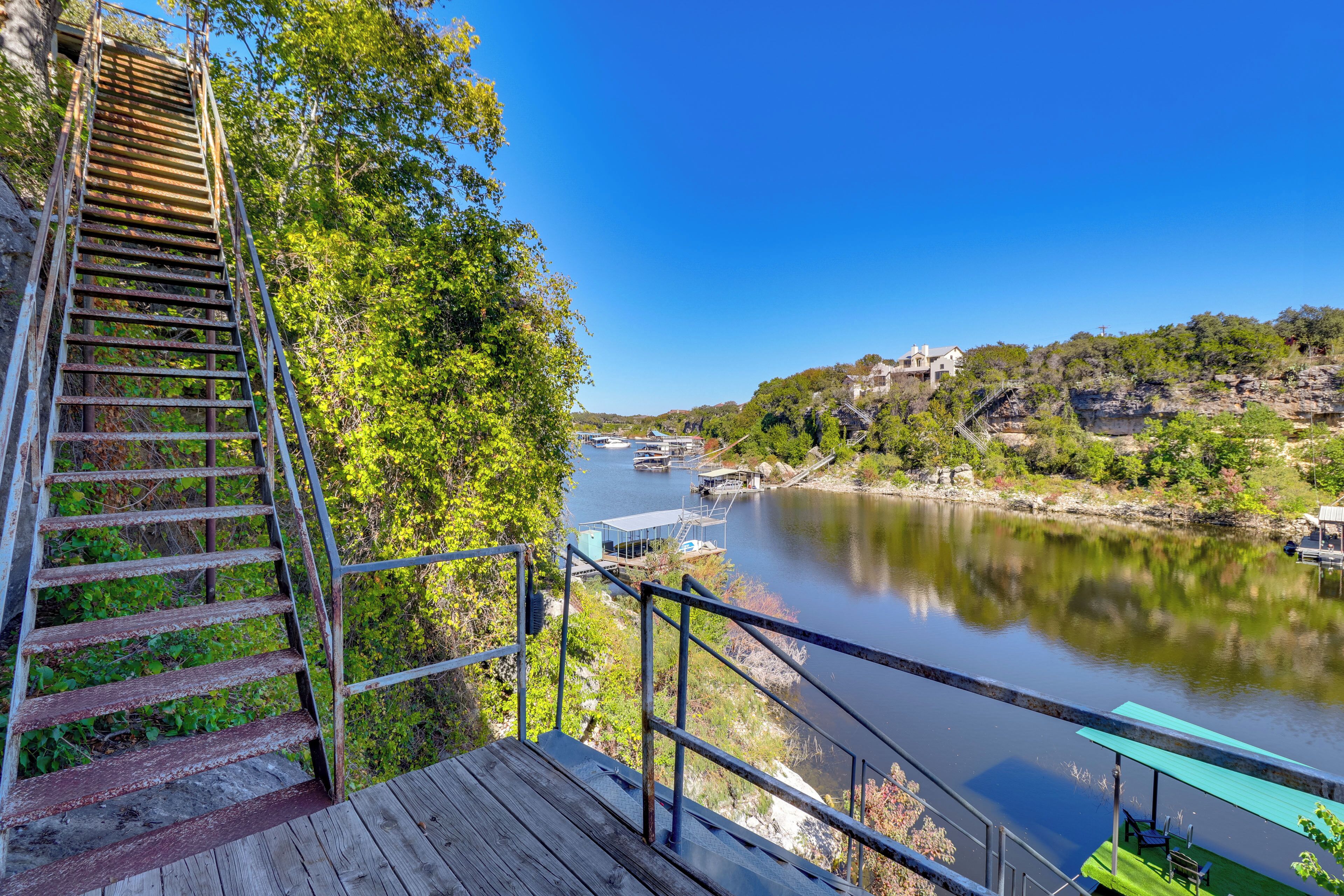 A wooden staircase leading to a viewing platform overlooking a calm river, surrounded by lush greenery and a clear blue sky.