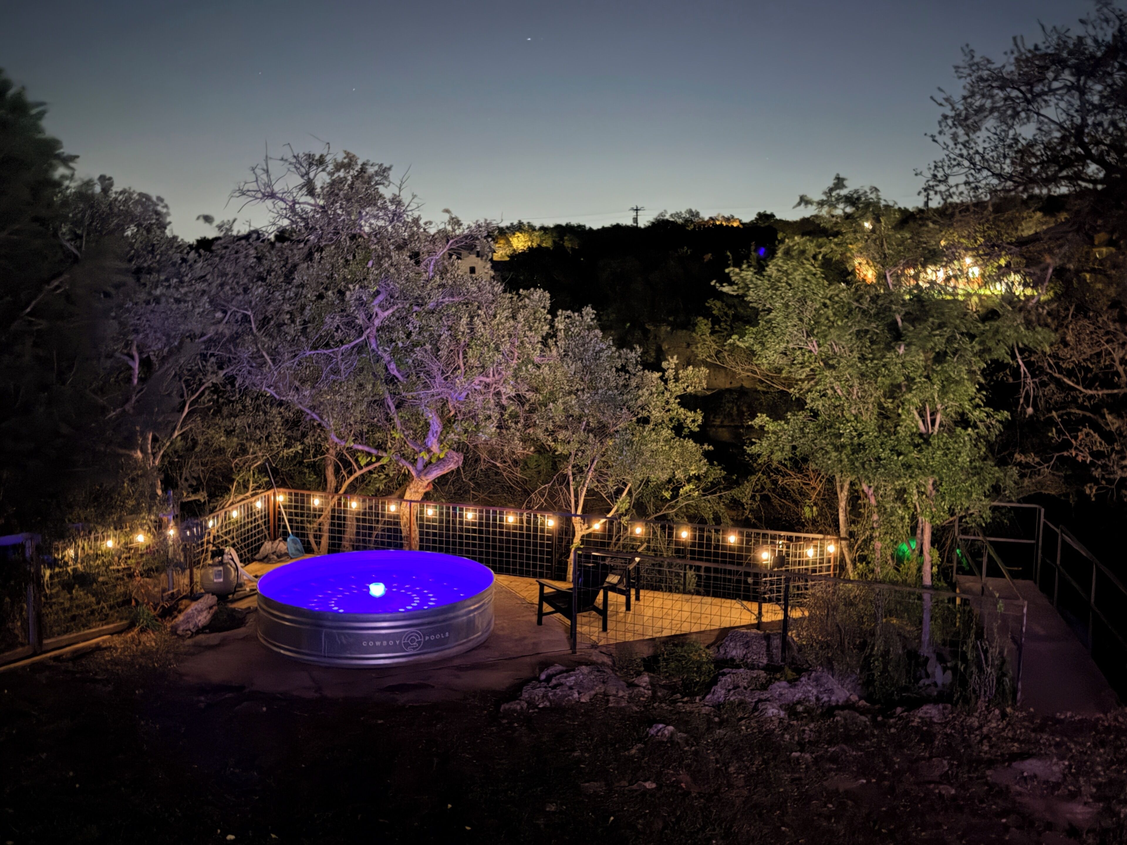 A circular pool with a blue light, surrounded by a fence and trees, in a nighttime setting.