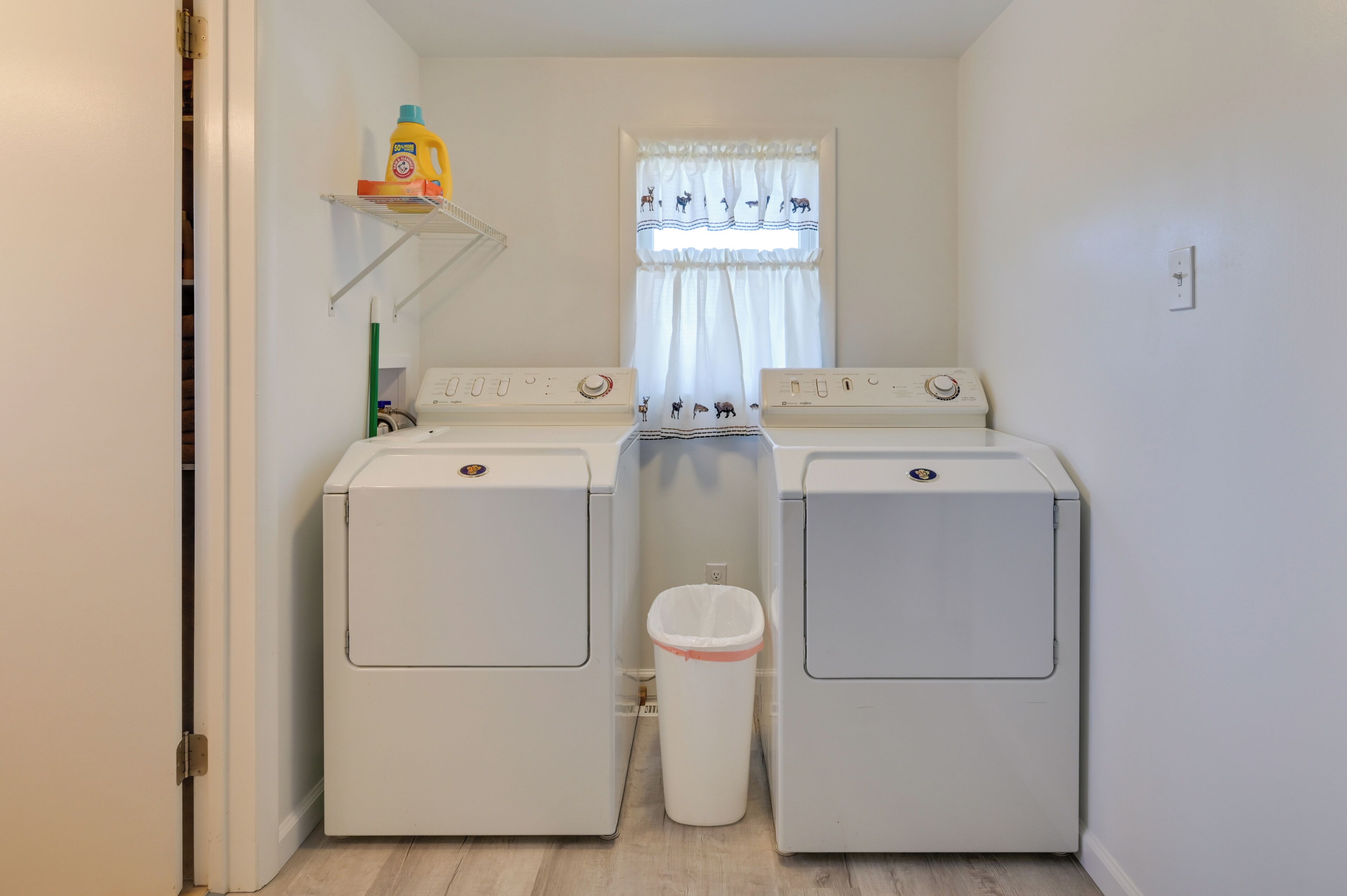 A laundry room with two front-loading washing machines and a dryer, a shelf with cleaning supplies, and a window with curtains.