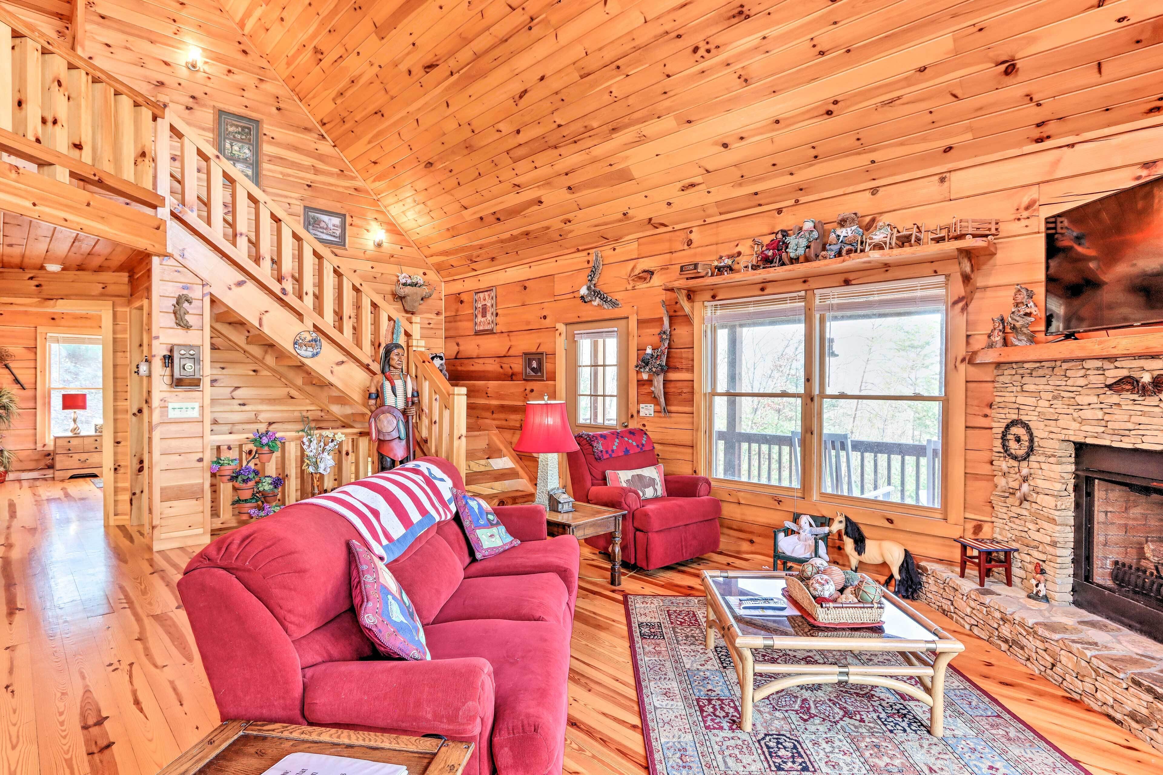 A log cabin interior with a red sofa, a fireplace, a staircase, and a large window.