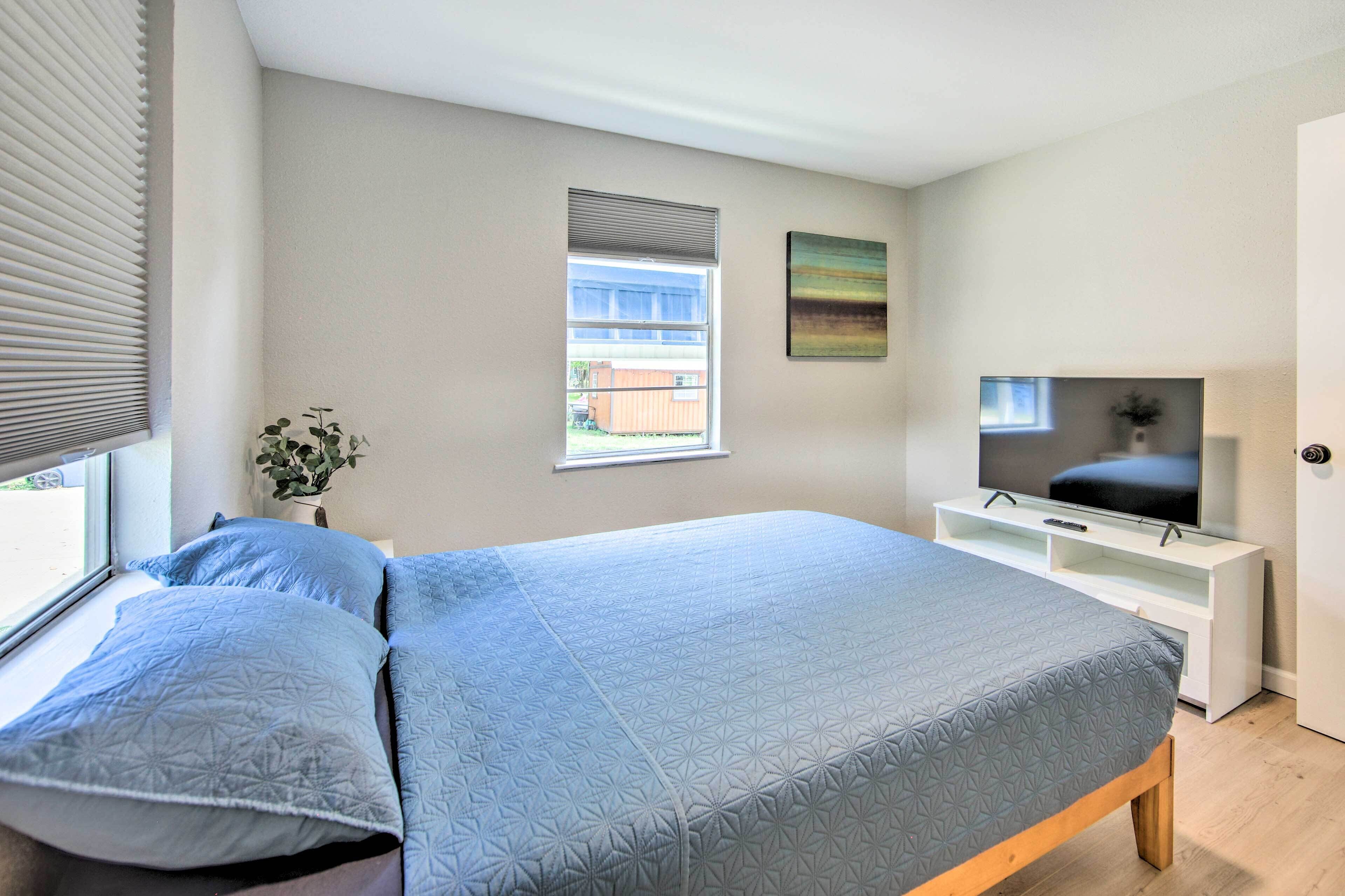 Une chambre à coucher avec un lit, un téléviseur posé sur un meuble, une fenêtre avec des stores et une plante.