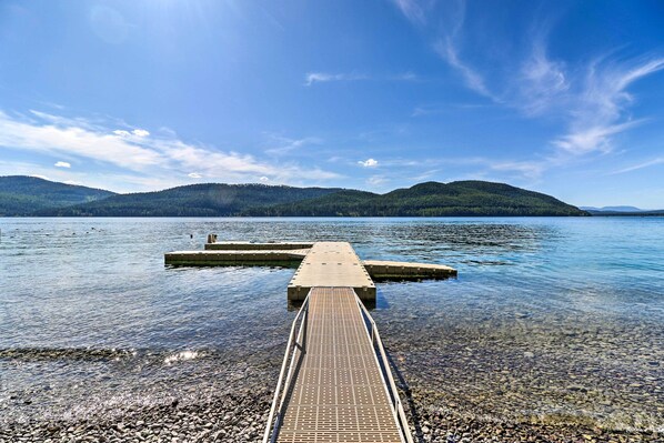 A clear view of a wooden dock extending into calm water, surrounded by a rocky shoreline and distant green hills under a blue sky.