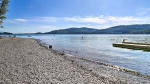 A pebbled beach with a dock extending into calm waters, surrounded by hills. - Whitefish Escape w/ Deck: 3 Mi to Ski Resort! (Whitefish)