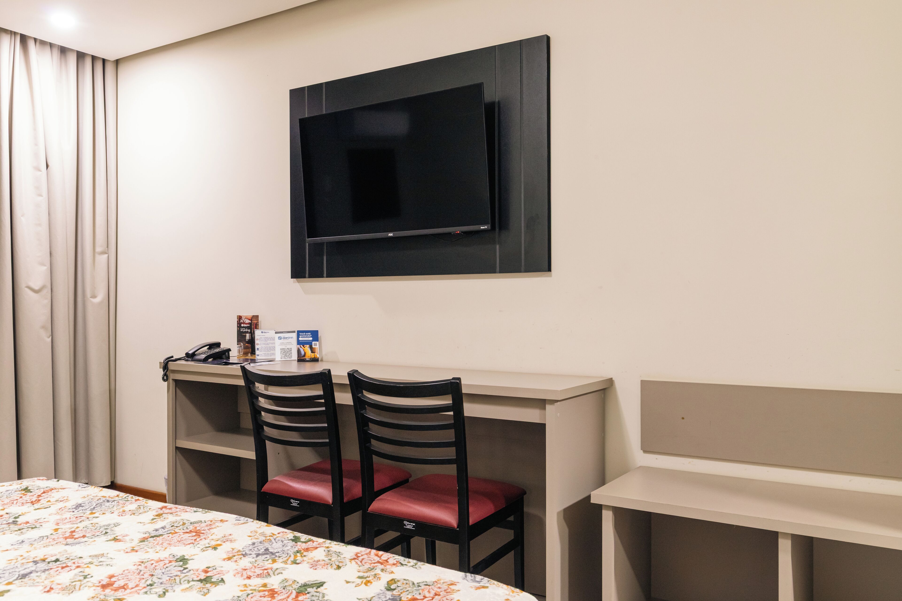 Habitación de hotel con cama, escritorio y dos sillas, televisor montado en la pared y ventana con cortinas.