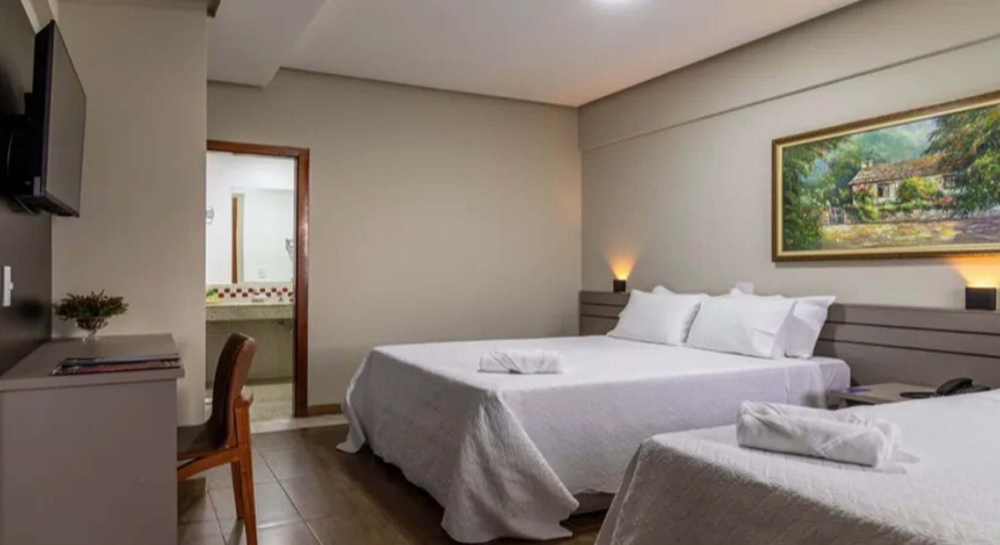Une chambre d’hôtel avec deux lits, un bureau, une chaise, un tableau au mur et une vue sur une maison par la fenêtre.