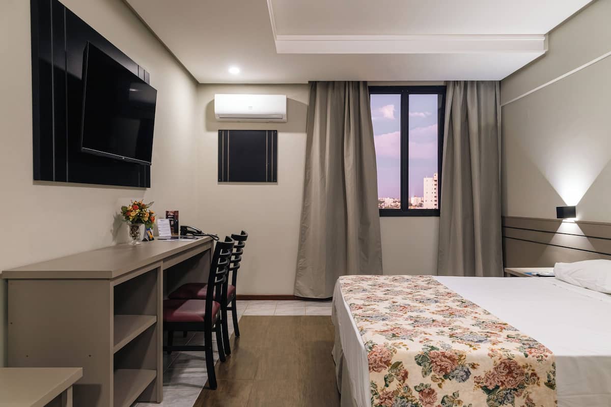 Hotelkamer met een bed, een bureau met stoel, een aan de muur bevestigde televisie, een airconditioning en een raam met gordijnen, met uitzicht op de stad.