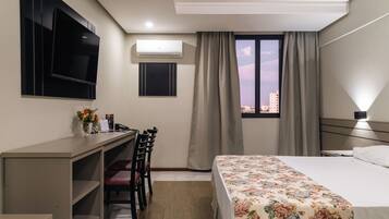 Une chambre d’hôtel comprenant un lit, un bureau avec une chaise, un téléviseur fixé au mur, un appareil de climatisation, une fenêtre avec des rideaux et une vue sur le paysage urbain.