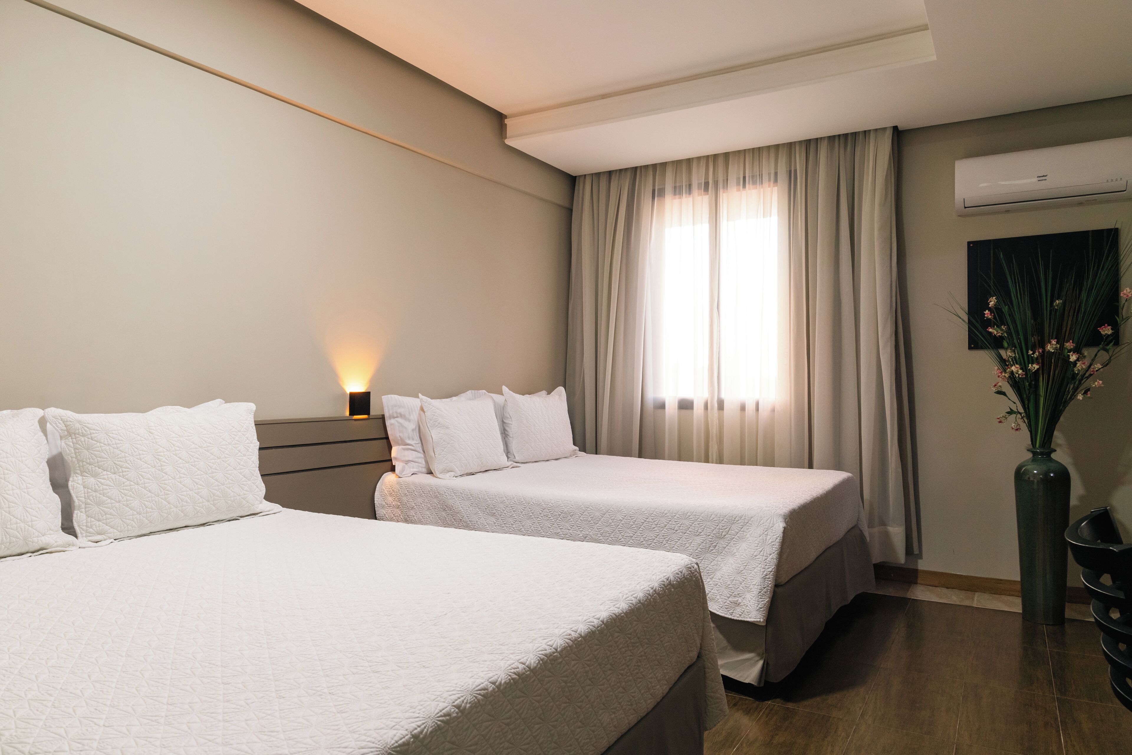 A hotel room with two beds, a nightstand, a vase with flowers, and a window with curtains.