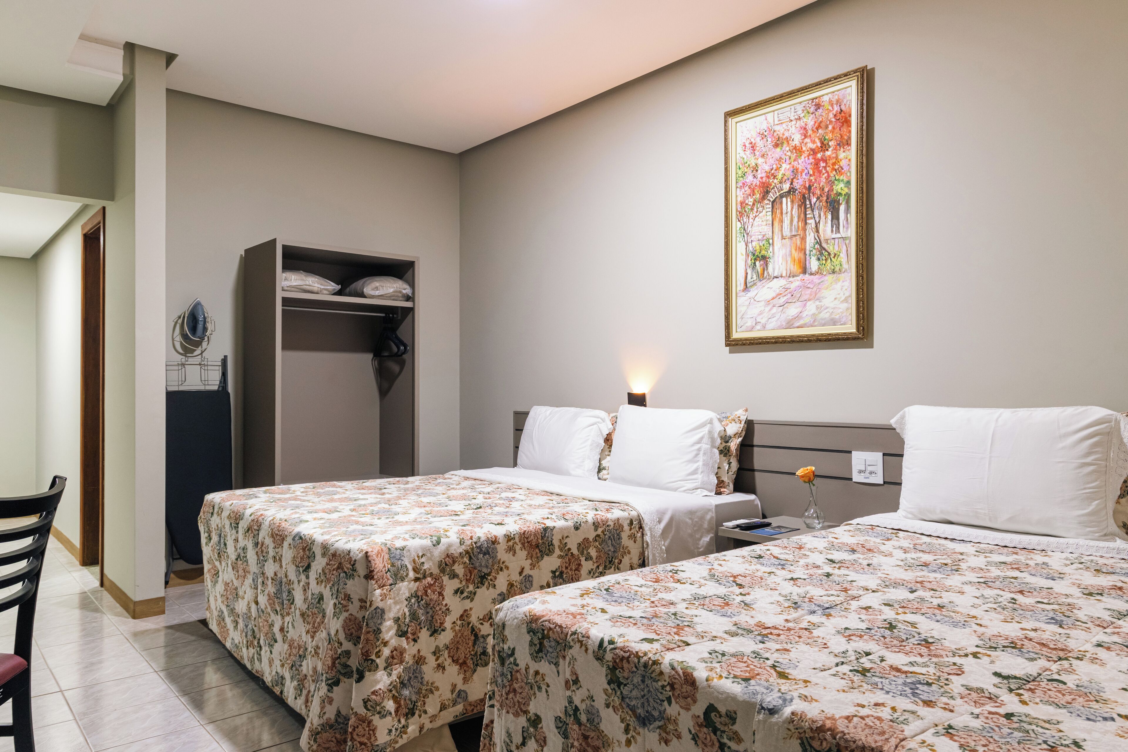 A hotel room with two beds, a wardrobe, a painting, and a desk with a chair.