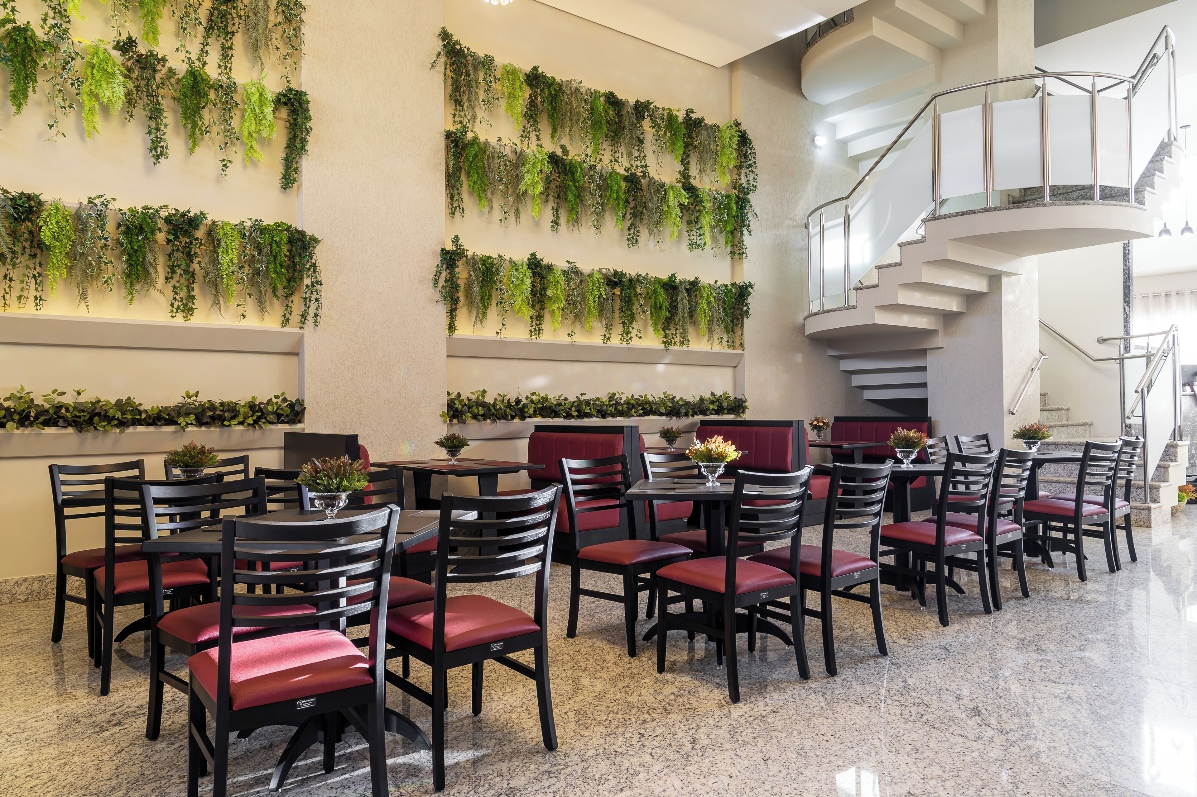 A hotel lobby with multiple dining tables and chairs, a staircase, and green plants on the walls.