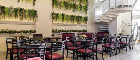 Un vestíbulo de hotel con varias mesas y sillas para comer, una escalera y plantas verdes en las paredes.