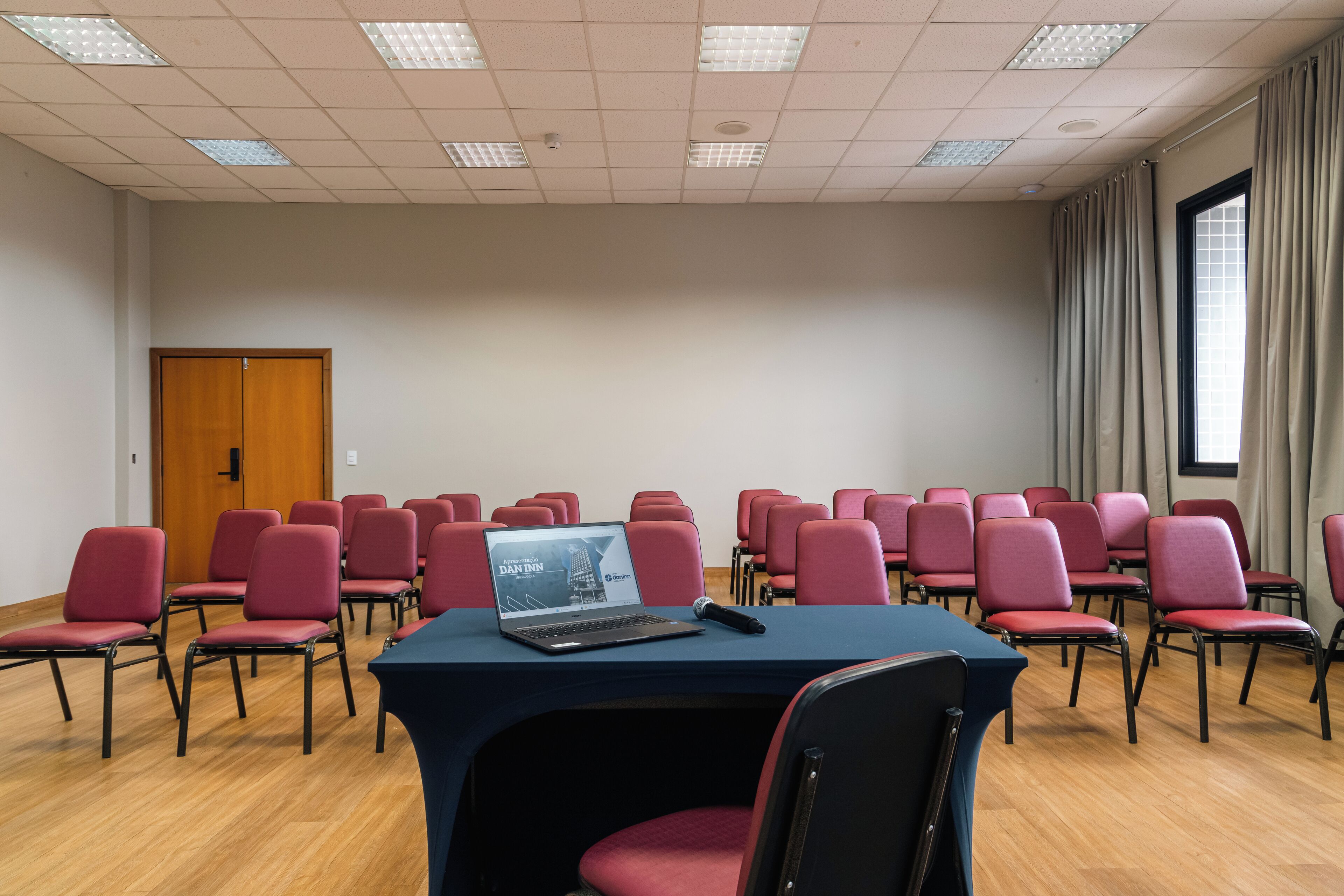 Une salle de conférence avec des rangées de chaises rouges, un ordinateur portable posé sur une table et une porte en bois.