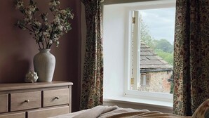 Ein Schlafzimmer mit einem Bett, einer Kommode, einer Vase mit Blumen und einem Fenster mit Vorhängen.